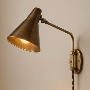 May include: A wall-mounted lamp with an antique brass finish, featuring a conical shade and an adjustable arm. The fixture includes a circular wall plate and a twisted fabric cord extending downward, providing a warm light from the bulb inside.