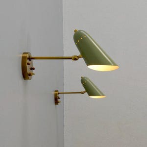 Op de afbeelding: Twee wandkandelaars in een mid-century modern design. Ze hebben olijfgroene conische kappen met kleine perforaties, messing armen en montageplaten. De lampen zijn op verschillende hoogtes aan een witte muur bevestigd.
