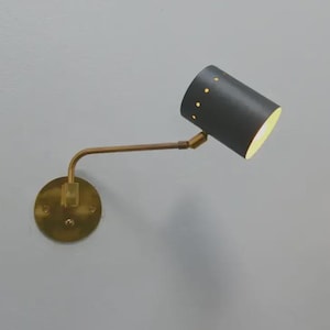 Può includere: Una lampada da parete con un paralume cilindrico nero e un braccio e una base color ottone. Il paralume presenta piccoli fori distanziati. La lampada è montata su una parete grigia, offrendo un'estetica moderna e minimalista.