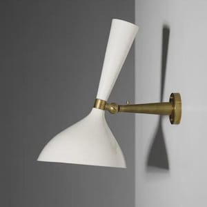 Könnte beinhalten: Eine weiße Wandleuchte im Mid-Century-Modern-Stil mit einem Messingarm und einer Montageplatte. Der Lampenschirm hat eine gebogene, konische Form. Die Leuchte ist an einer hellgrauen Wand montiert.