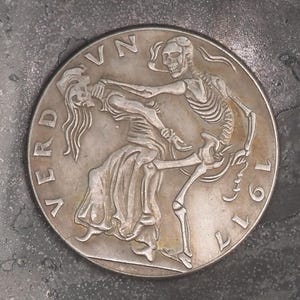 Può includere: Una moneta d'argento con un disegno in rilievo di uno scheletro che abbraccia una donna. La moneta reca l'iscrizione "VERD" e l'anno "1911". La moneta è posta su uno sfondo scuro e strutturato.