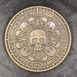 Könnte beinhalten: Eine goldfarbene Münze mit einem Totenkopf- und Knochenkreuz-Design. Die Münze trägt die Inschriften "Memento Mori" und "Remember You Must Die" am Rand.