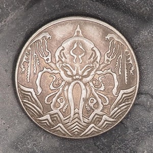 Könnte beinhalten: Eine silberne Münze mit einer detaillierten Gravur eines Cthulhu-ähnlichen Wesens mit Tentakeln. Die Münze befindet sich auf einem dunkelgrauen Hintergrund.