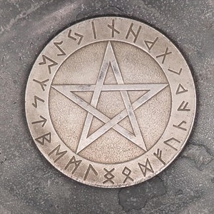 May include: A silver-coloured metal pentagram with a circle of runes around the outside. The pentagram is on a grey textured surface.