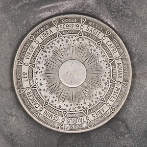 May include: A silver circular zodiac wheel with the names of the twelve zodiac signs in English. The wheel is surrounded by a ring of stars and has a sun in the center.