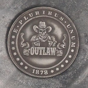 May include: A silver-colored coin with a skull wearing a cowboy hat and the words "E. Pluribus Unum" and "Outlaw" on it. The coin also has the year "1878" on it.