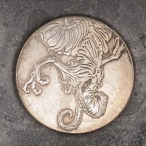 May include: A silver coin featuring an intricate engraving of a mythical creature with wings and a long tail. The creature is shown in a dynamic pose, with its wings outstretched and its tail curled.