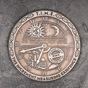 May include: A circular, silver-toned metal clock face with Roman numerals and the text "T.I.M.E. THE INCREMENT MEASURING EXISTENCE" around the edge. The clock face features a sun, moon, and a clock hand pointing to the number 5. The text "CHAUTAUQUA'S TIME WORKS ONE OUNCE 1994 - BE SELVES" is also visible on the clock face.