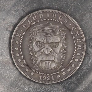 May include: A silver coin featuring a monkey's face in the centre. The coin is inscribed with the words "E. Pluribus Unum" around the edge and the year "1921" at the bottom.