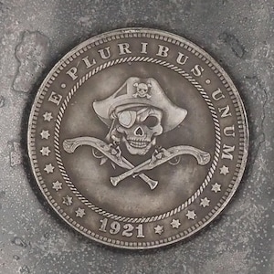 Pode incluir: Uma moeda de prata com um desenho de caveira e ossos cruzados. A moeda tem as palavras "E. Pluribus Unum" e "1921" inscritas nela.