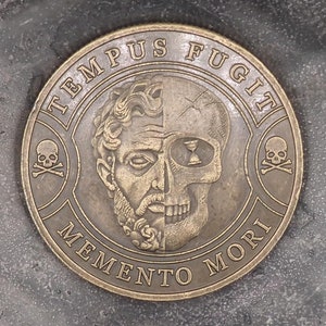Puede incluir: Una moneda redonda de color dorado con la frase latina "Tempus Fugit Memento Mori" y un diseño de calavera y huesos cruzados.