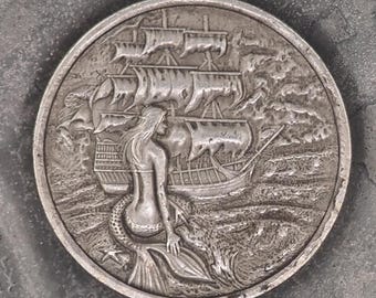 Puede incluir: Una moneda de plata con un diseño en relieve de una sirena sentada en la proa de un barco. El barco navega en un mar ondulado.
