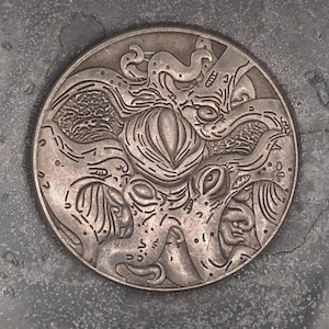 Könnte beinhalten: Eine silberne Münze mit einem komplizierten Design einer Kreatur mit mehreren Tentakeln. Die Münze hat einen erhöhten Rand und eine glatte Oberfläche.