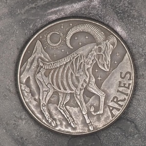 May include: A silver coin with a skeletal ram and the word "ARIES" inscribed around the edge. The coin has a crescent moon and stars in the background.