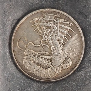 Puede incluir: Moneda de plata con una imagen detallada de una cobra con una corona en la cabeza. La moneda tiene la inscripción "The Egyptian".