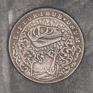 Pode incluir: Uma moeda de prata com um design detalhado que apresenta um crânio de dinossauro e as palavras "E Pluribus Unum".