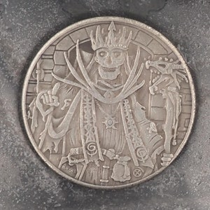 Puede incluir: Una moneda de plata con un diseño detallado de una figura esquelética que lleva una corona y una túnica, sosteniendo un bastón y un dragón. La moneda tiene una superficie texturizada y un borde elevado.