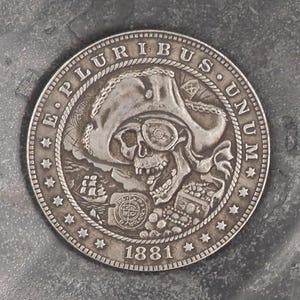 Könnte beinhalten: Eine Silberdollar-Münze mit einem Totenkopf, der einen Piratenhut und eine Augenklappe trägt. Die Münze trägt die Inschriften "E PLURIBUS UNUM" und "1881".