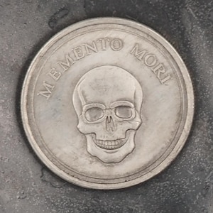 May include: A silver coin featuring a skull and the inscription "MEMENTO MORI". The coin is set against a dark, textured surface. The skull's features are clearly defined, with detailed eye sockets and a smiling expression.
