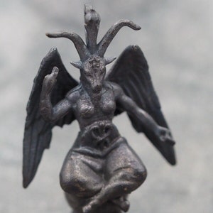 Może przedstawiać: Czarna figurka demona z trzema rogami, skrzydłami i głową kozła. Demon siedzi w pozycji medytacyjnej na kwadratowej podstawie.
