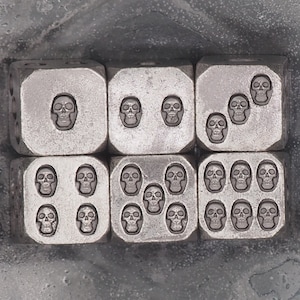 May include: Six dice with skull symbols in a square arrangement. The dice are a metallic gray color and have a skull design for each number. The background is a textured, gray surface.