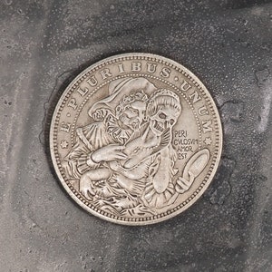 May include: A silver coin featuring the inscription "E Pluribus Unum" and a design of two figures, one holding a mirror. The coin is resting on a grey textured surface.