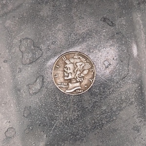 May include: A silver-coloured coin with a profile of a woman's face on it. The coin is on a grey, textured surface. The coin says "IN GOD WE TRUST" and "1943" on it.