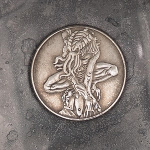 May include: A silver coin featuring a detailed image of a creature with tentacles and a human-like body.