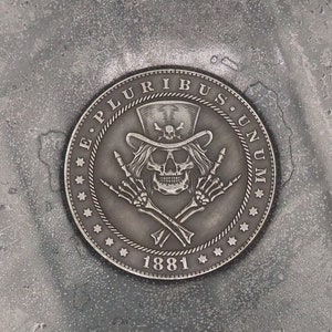 Op de afbeelding: Een zilveren munt met een schedel die een hoge hoed draagt en de rock-and-roll handbeweging maakt. De munt heeft de woorden "E. PLURIBUS UNUM" en "1881" erop gegraveerd.