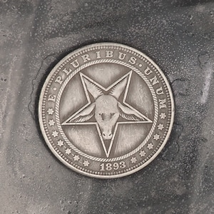 May include: A silver coin with a pentagram and a goat's head in the center. The words "E. PLURIBUS UNUM" are inscribed around the edge of the coin, and the year "1893" is inscribed at the bottom.