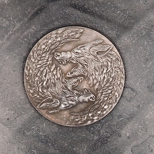 May include: A silver-toned metal coin with a design of two wolves facing each other. The wolves are in a circular design with their tails intertwined. The coin is on a gray textured surface.