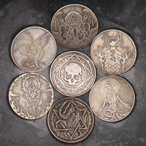 May include: Seven silver-toned metal tokens with various designs, including a skull, a creature, and a tree, arranged in a circle in a black bowl.