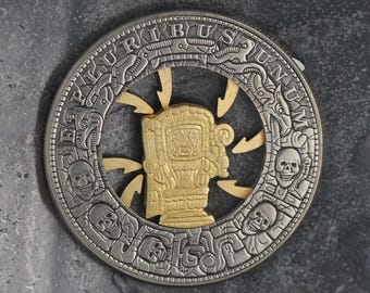 May include: A silver coin with a gold-colored design in the center. The coin features a circular design with the words "PLURIBUS UNUM" around the edge. The center design is a stylized figure with a skull-like face.