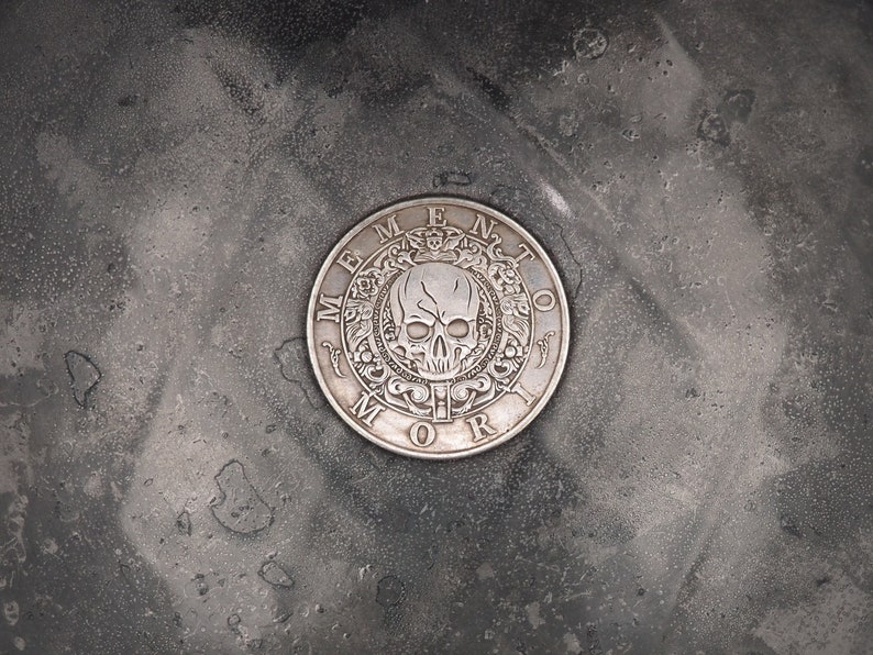 May include: A silver-colored round coin with a skull and crossbones design and the words "Memento Mori" around the edge.