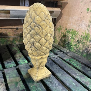 Puede incluir: Una escultura de piña de pino de hormigón amarillo con una superficie detallada y texturizada. La escultura está sobre una plataforma de madera.