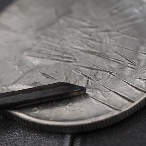 Op de afbeelding: Een close-up van een zilveren munt met een ontwerp van stralende lijnen. Een zwart metalen gereedschap rust op de munt.