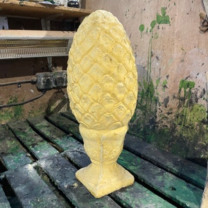 Puede incluir: Un remate de piña de cerámica amarillo con una superficie texturizada detallada. El remate es un elemento decorativo para muebles u otros objetos.