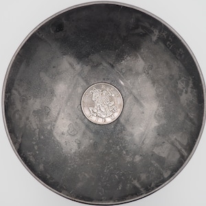 May include: A silver coin with a design of three figures and the text "CRAPE DIEU" is sitting in a gray metal bowl.