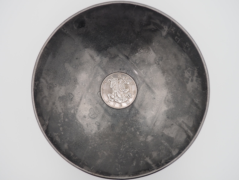 May include: A silver coin with a design of three figures and the text "CRAPE DIEU" is sitting in a gray metal bowl.