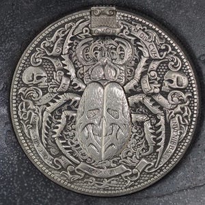 May include: A silver coin with a detailed design of a beetle with a skull on each side. The coin has the text "OPVS  OMNIVM  CARISS  IN  DIV  CIV  INV  ENIV  BAT  AMMXVII" around the edge.