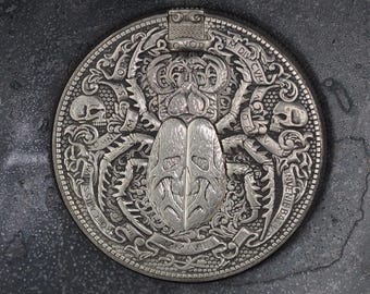 May include: A silver coin with a detailed design of a beetle with a skull on each side. The coin has the text "OPVS  OMNIVM  CARISS  IN  DIV  CIV  INV  ENIV  BAT  AMMXVII" around the edge.