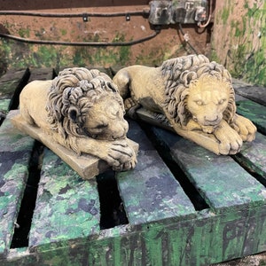 May include: Two concrete lion statues, each with a detailed mane and a sleeping pose. The lions are resting on a wooden surface with a green and brown camouflage pattern.