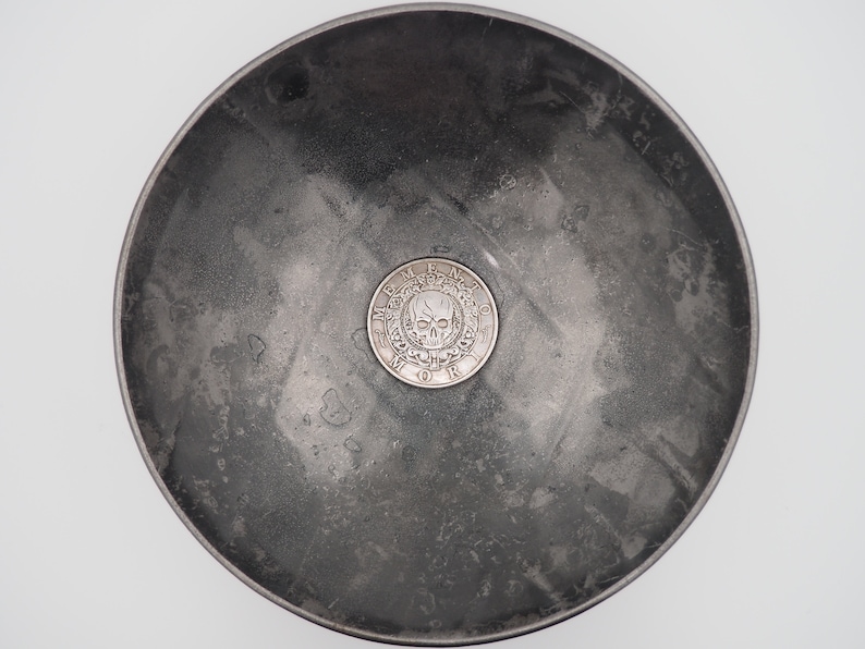 May include: A silver coin with a skull and crossbones design and the words "Memento Mori" around the edge, resting in a gray metal bowl.