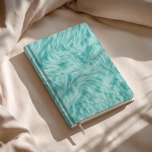 May include: A hardcover journal with a teal, faux fur cover. The notebook has a matching teal spine and a light pink ribbon bookmark. The journal is lying on a beige fabric surface.