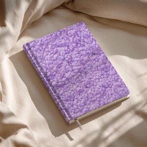 May include: A hardcover journal with a textured, light purple cover. The cover has a soft, plush appearance. A white ribbon bookmark is visible. The journal is lying on a beige surface.