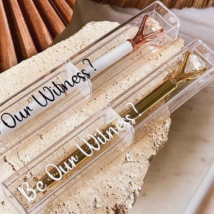 May include: Two pens in clear plastic cases, one with a gold diamond-shaped top and the other with a rose gold top. The pens are white with the text "Be Our Witness?" on the case. The pens are on a textured surface.