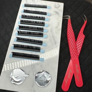 Op de afbeelding: Een wit marmeren dienblad met rijen zwarte wimperextensies die zijn gelabeld met metingen in millimeters. Twee rode pincetten met gouden letters bevinden zich aan de rechterkant van het dienblad. Twee zilveren folie cirkels bevinden zich aan de linkerkant van het dienblad.