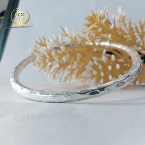 Puede incluir: Un brazalete de plata martillado. El brazalete es liso y brillante, con una superficie ligeramente texturizada. El brazalete está sobre una superficie blanca.
