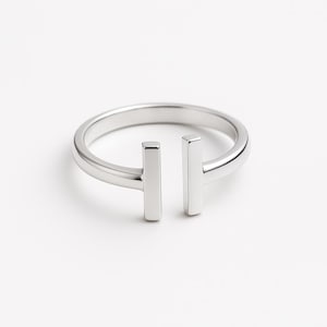 Op de afbeelding: Een minimalistische zilveren ring met een modern design. De ring heeft een cirkelvormige band met twee verticale rechthoekige staven, waardoor een unieke T-vorm ontstaat. De ring is gemaakt van glanzend, reflecterend metaal en staat tegen een witte achtergrond.