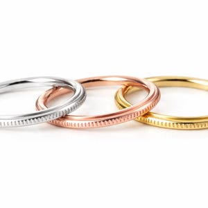 May include: Three thin rings in silver, rose gold, and gold. Each ring has a textured, dotted pattern around the band. The rings are displayed on a white background, showcasing their different metallic colors.